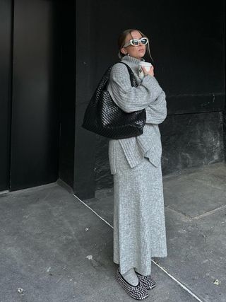 a photo of an influencer wearing a gray turtleneck top with a matching maxi skirt and socks with rhinestone ballet flats and a woven shoulder bag