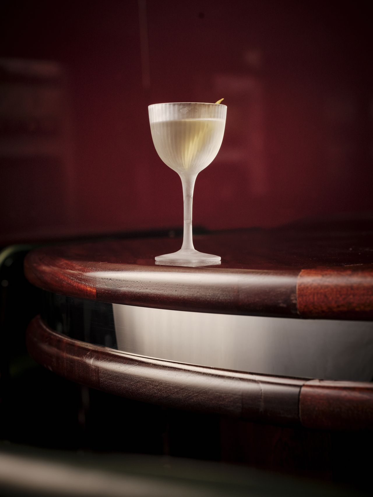 A matte, semi-transparent glass filled with an equally transparent drink and a slice of lemon sits atop a wooden table with a textural look.