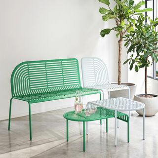 Image of a metal green and light blue garden bench and matching nesting table. They are staged in a room with white walls, concrete floors, and two potted trees.