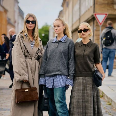 Women pose for a picture. One wears a trench coat, the middle one wears a sweater layered over a button down with jeans , and the last one wears a pleated skirt and short-sleeve sweater. 