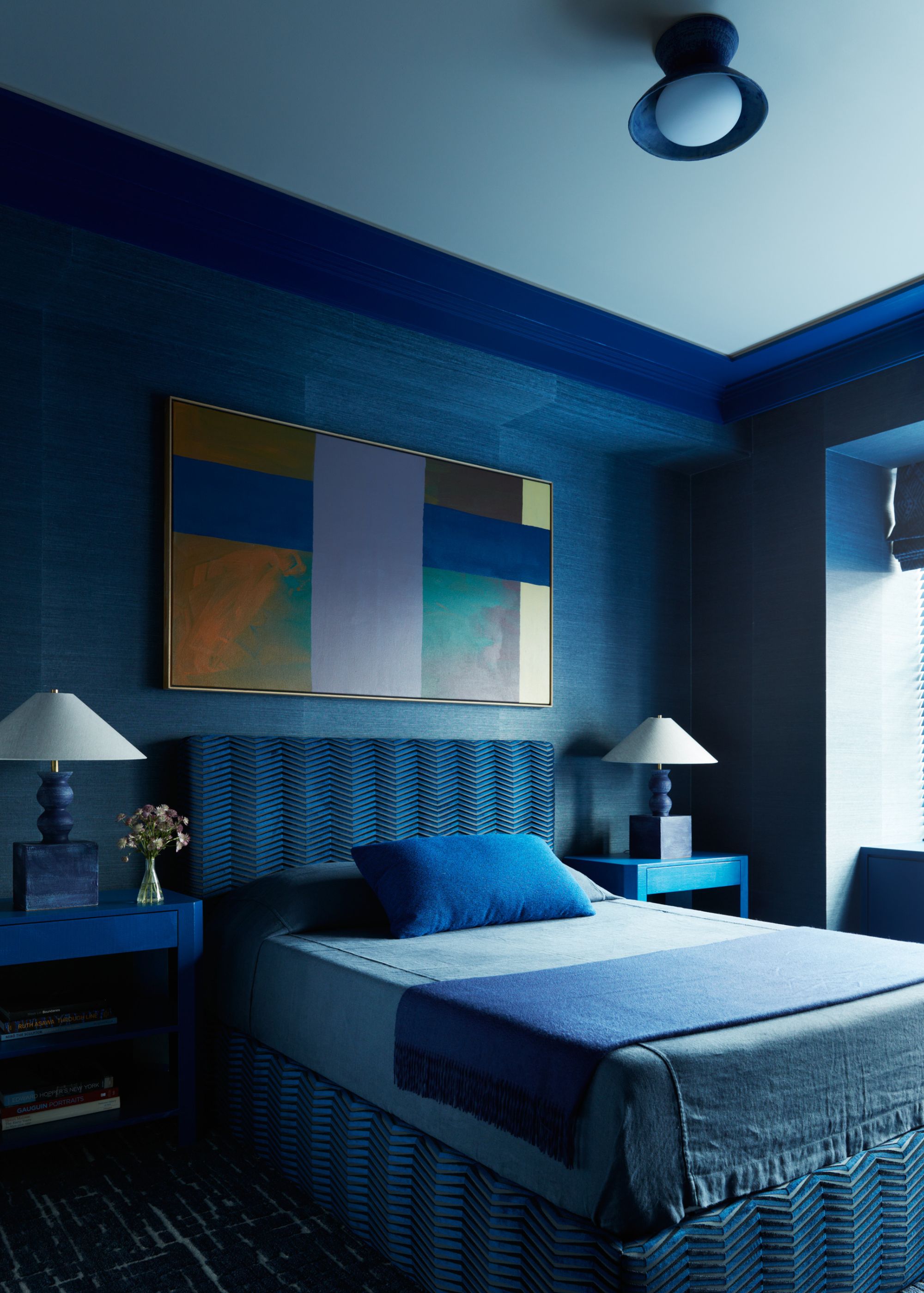 A modern bedroom with a dark blue wallcovering on the walls, dark blue paint on the trim, a pale blue ceiling, and a bed with a dark blue headboard and minimalist blue bedding. A block-pattern abstract artwork on the walls in a palette of lilac, blue, and orange.