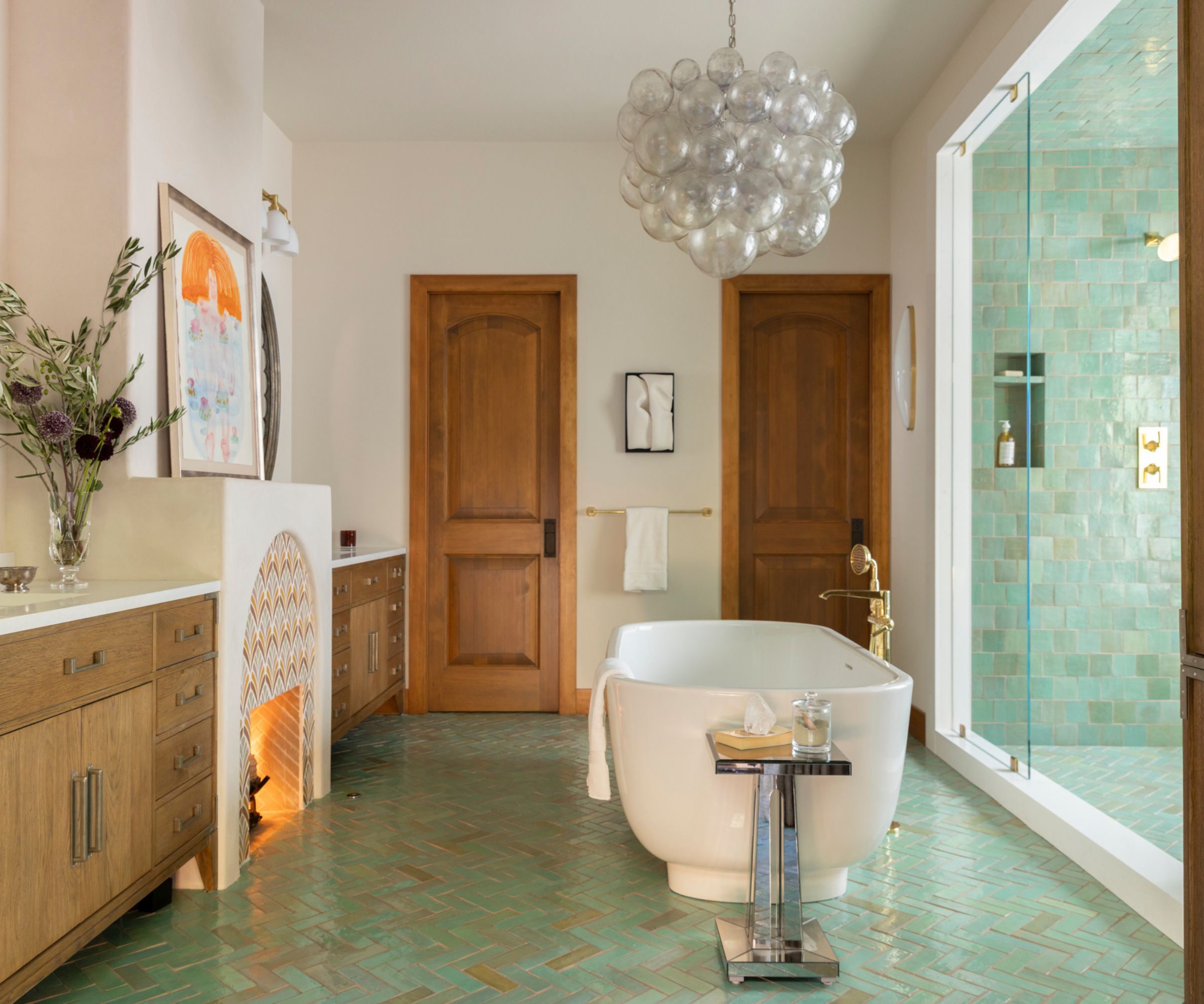 A large bathroom with two wooden vanities flanking a statement fireplace, blue floor tiles, and a freestanding bath