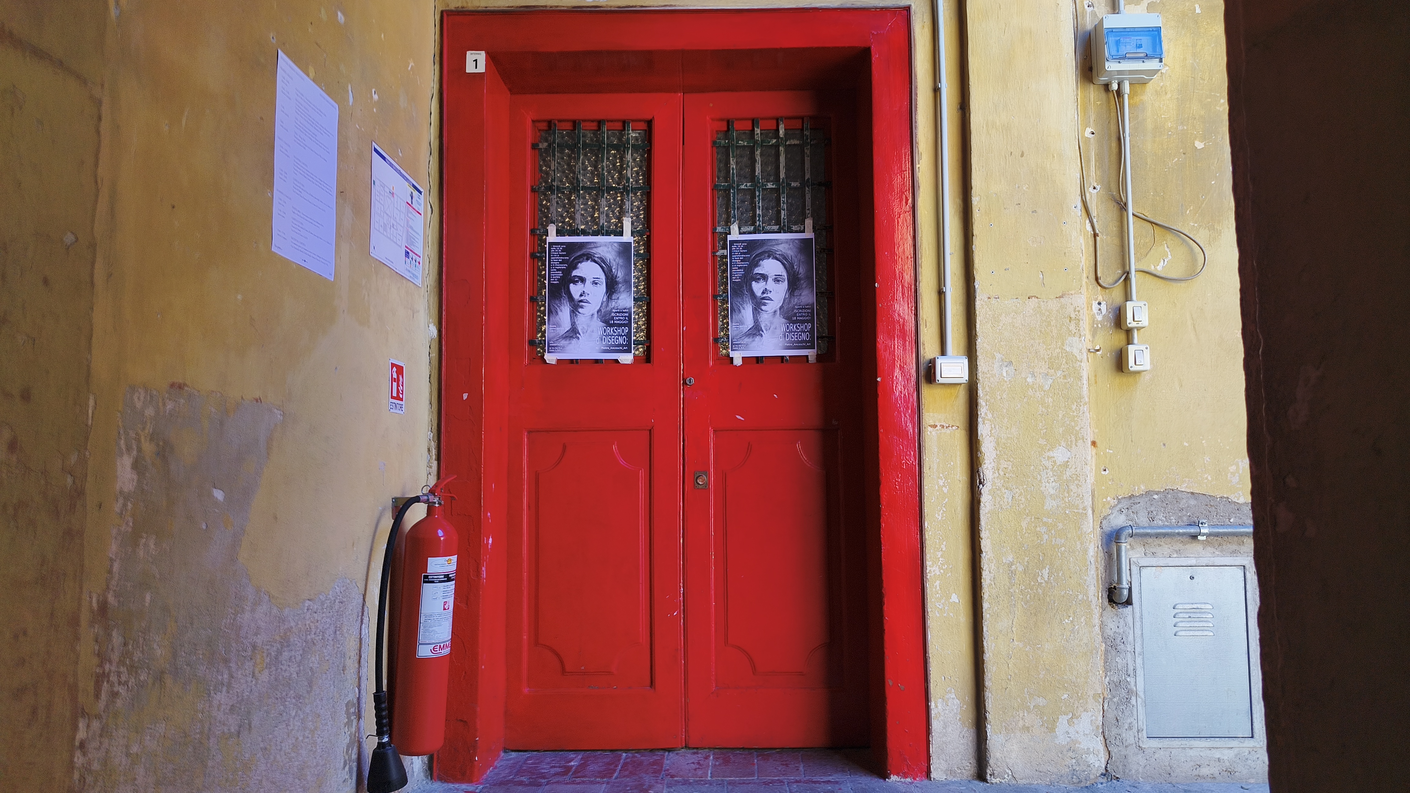A lone visitor, silhouetted against a red doorway at the top of a narrow stone staircase, with a sequence of black-and-white photographs pinned along the left-hand wall.