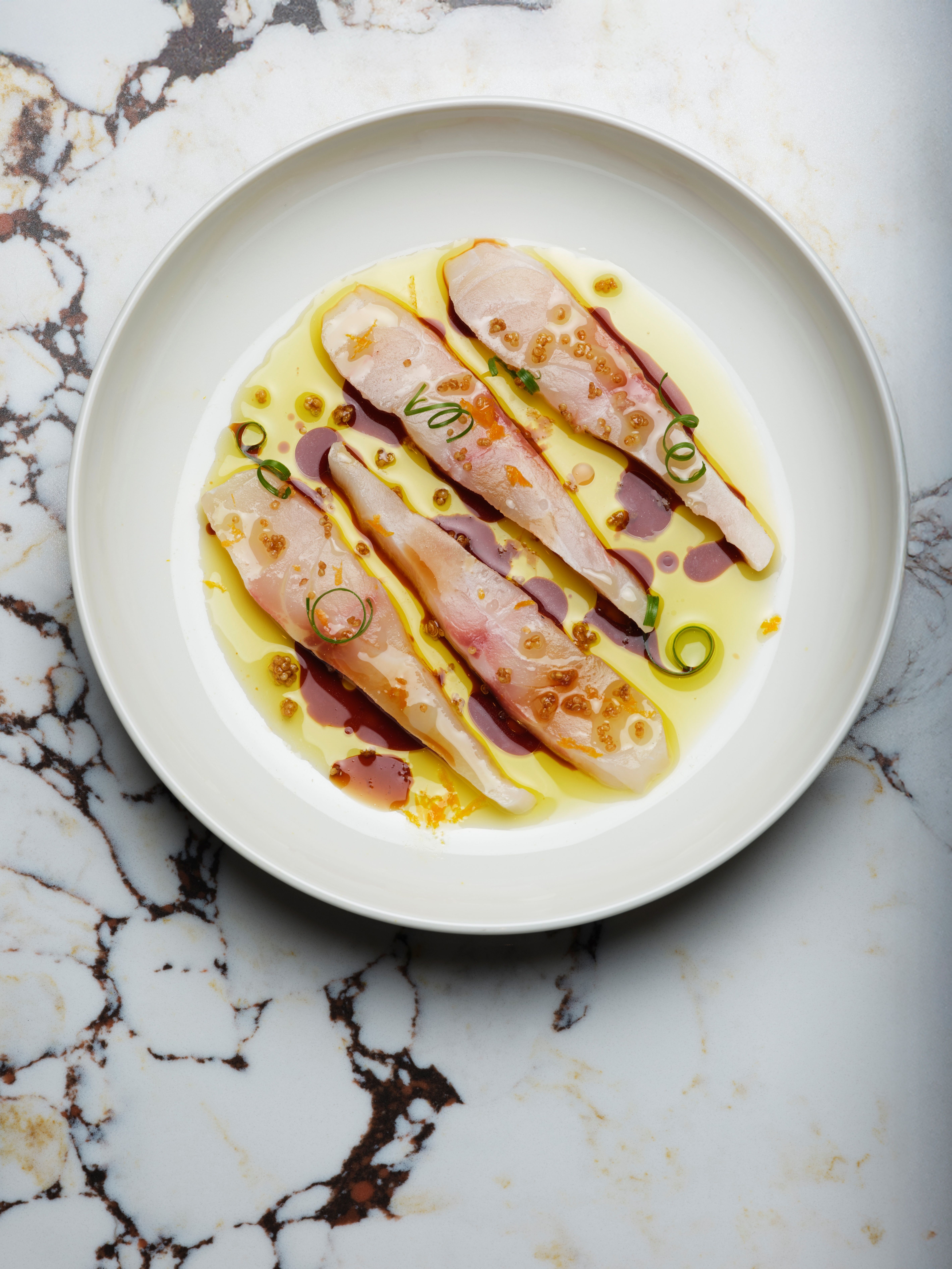 A white ceramic plate with seabass crudo, sprinkled with oil, baslamic vinegar, and spring onions, sits atop a white and black marble top, while in another image, a bottle of red wine sits on a wooden table next a black ceramic snack plate topped with meat, cherry, and cheese skewers.