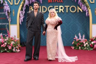 From left to right: Luke Newton in a grey suit and Nicola Coughlan in a pink dress and maroon gloves holding hands on the capret at a screening of Bridgerton Season 3 in London.