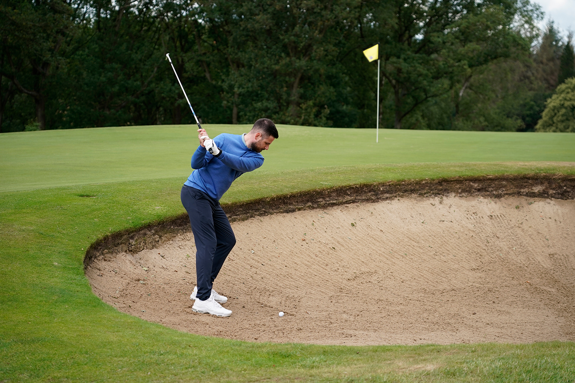 Baz Plummer at the top of backswing in the bunker, attempting to play a bunker shot towards a middle flag on the 8th green at Sand Moor Golf Club