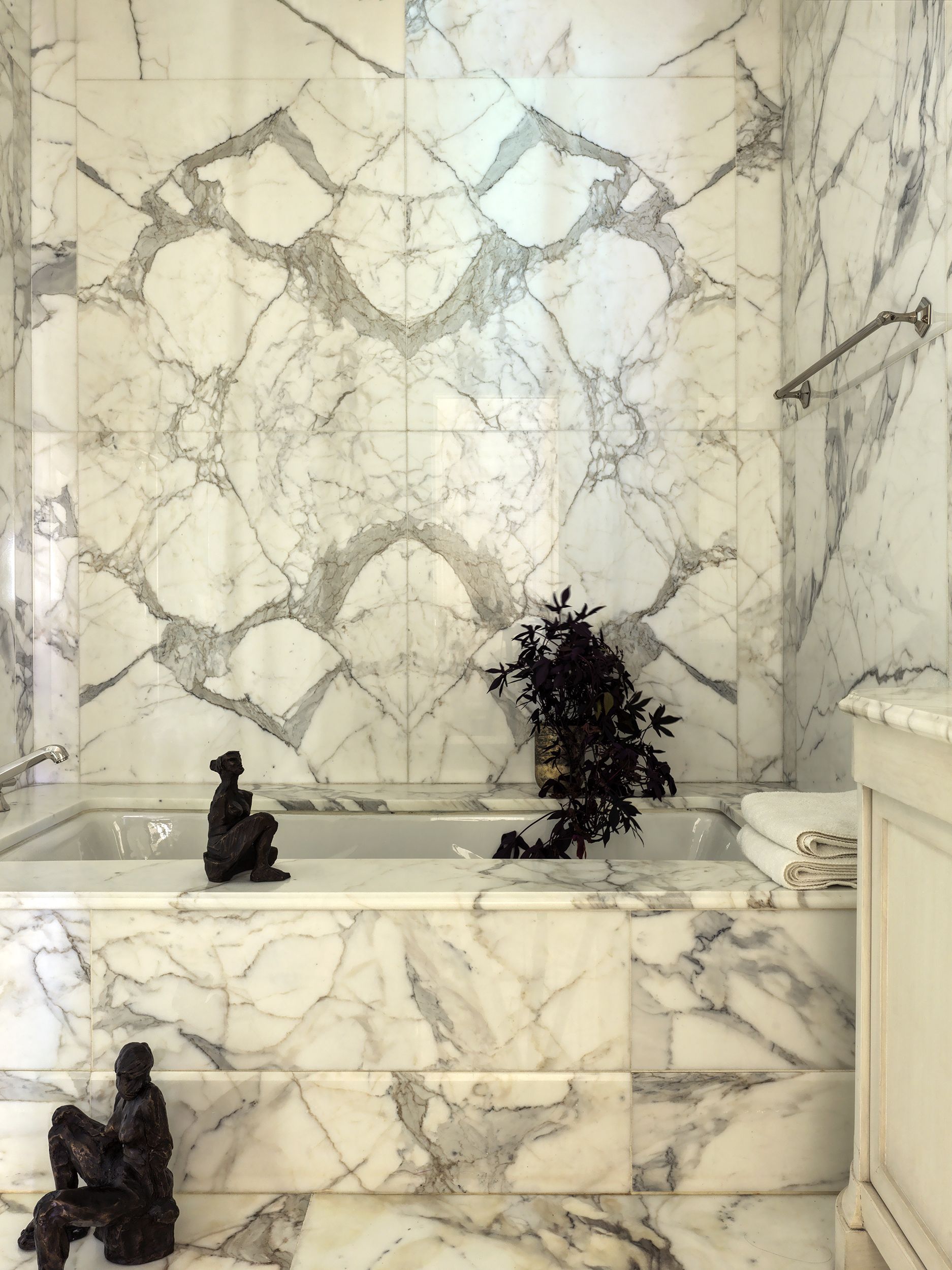 A white and gray-veined marble-drenched bathroom with a black trailing plant by the bath with two black decorative figures
