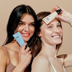 Two women holding Eight Saints Skincare products near their faces while posing against a beige background