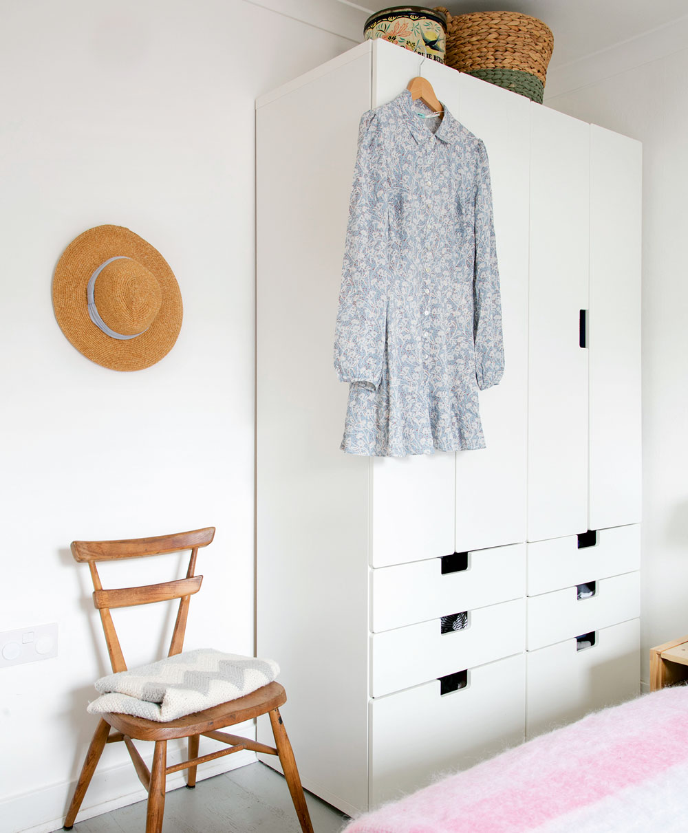 Bedroom with white wardrobe with storage baskets on top next to wooden chair.