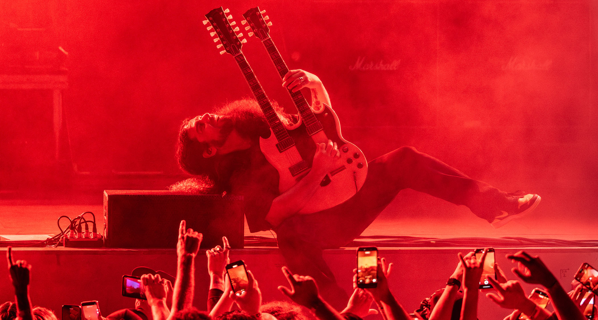 Claudio Sanchez throws a move on stage as he plays his white doubleneck. The stage is lit in deep red.