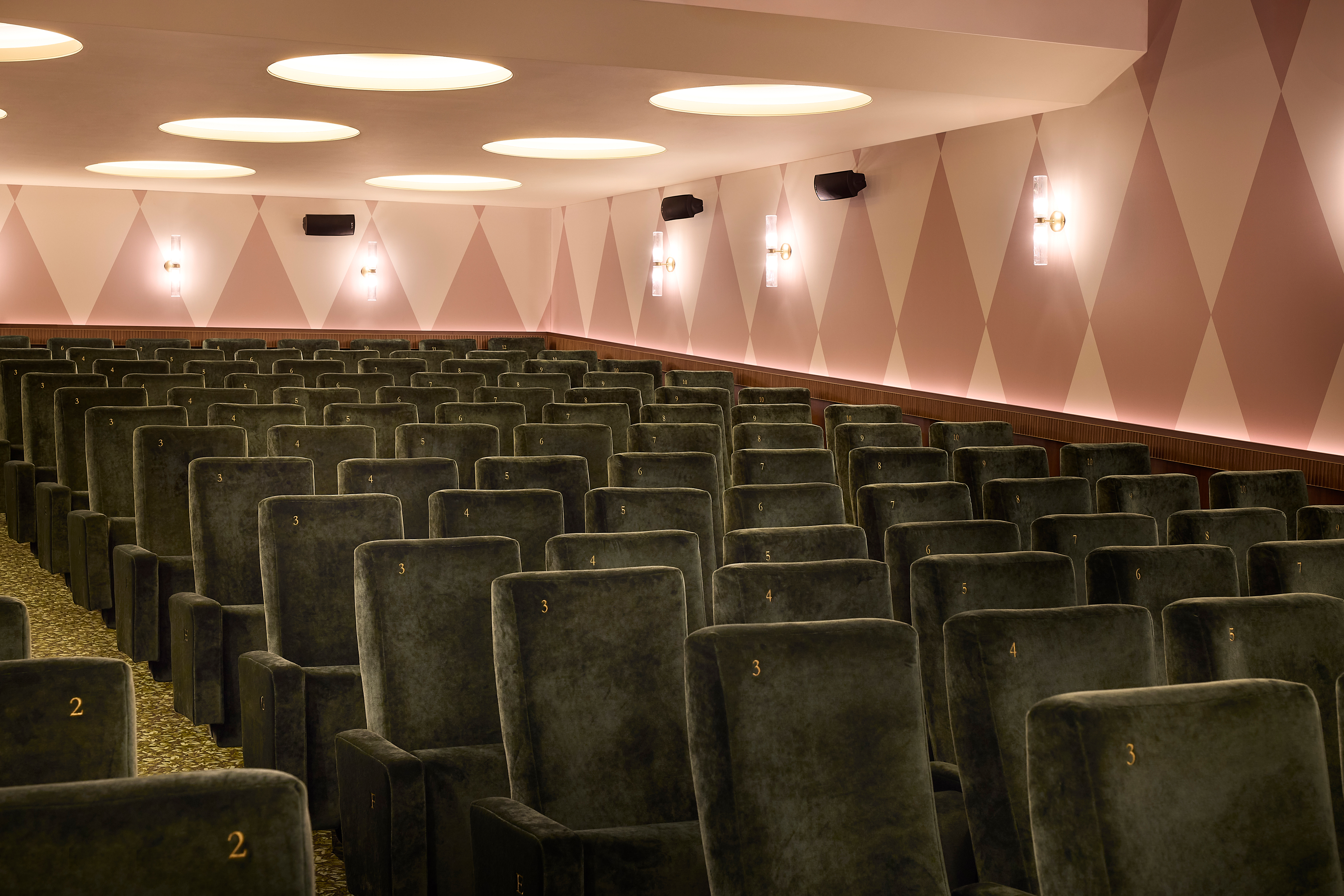 A series of interior shots depicting the retro-futuristic decor of a cinema captures velvet-clad rooms with a palette of reds, pinks, and pale dark greens, theatrical doorways, either arched or draped in curtains, ceiling porthole windows, and a vintage, golden entrance with red neon lights.