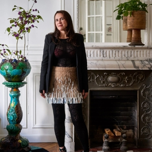 Image of a woman in a black and tan outfit standing in front of the fireplace in a maximalist living room.