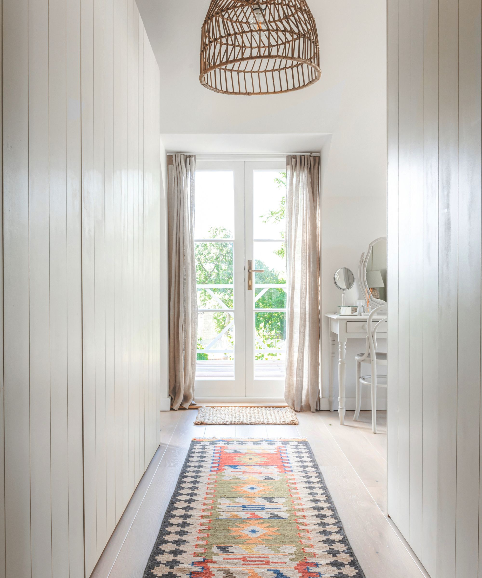 A white painted minimalist hallway with a bright rug, leading towards glass double doors to a garden.