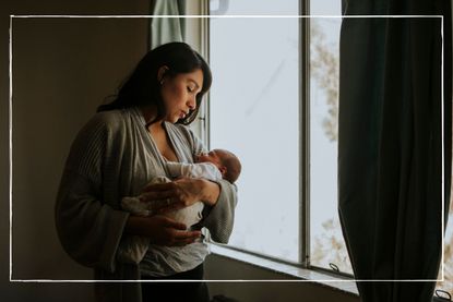 mother holding newborn baby in front of a window at home