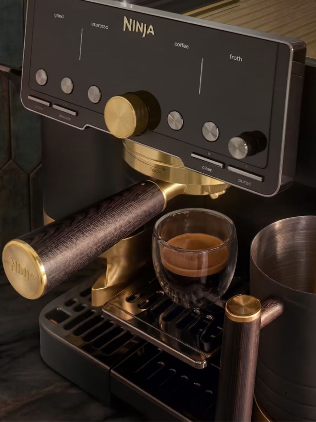 Close-up image of a dark gray and gold espresso machine with a wooden and gold portafilter brewing an espresso shot into a glass espresso cup.