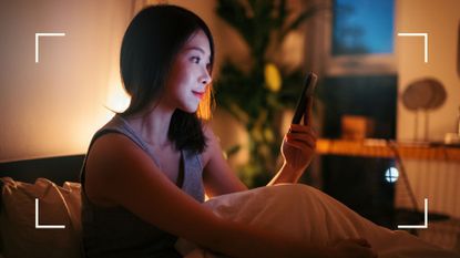 Woman looking at phone in bed under dimmed lighting, researching can you burn fat while you sleep