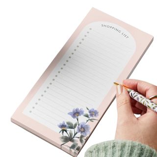 A hand with a sage green wooly jumper sleeve holding a floral white and gold pen over a pale pink and white shopping list with purple flower detailing in the bottom right corner