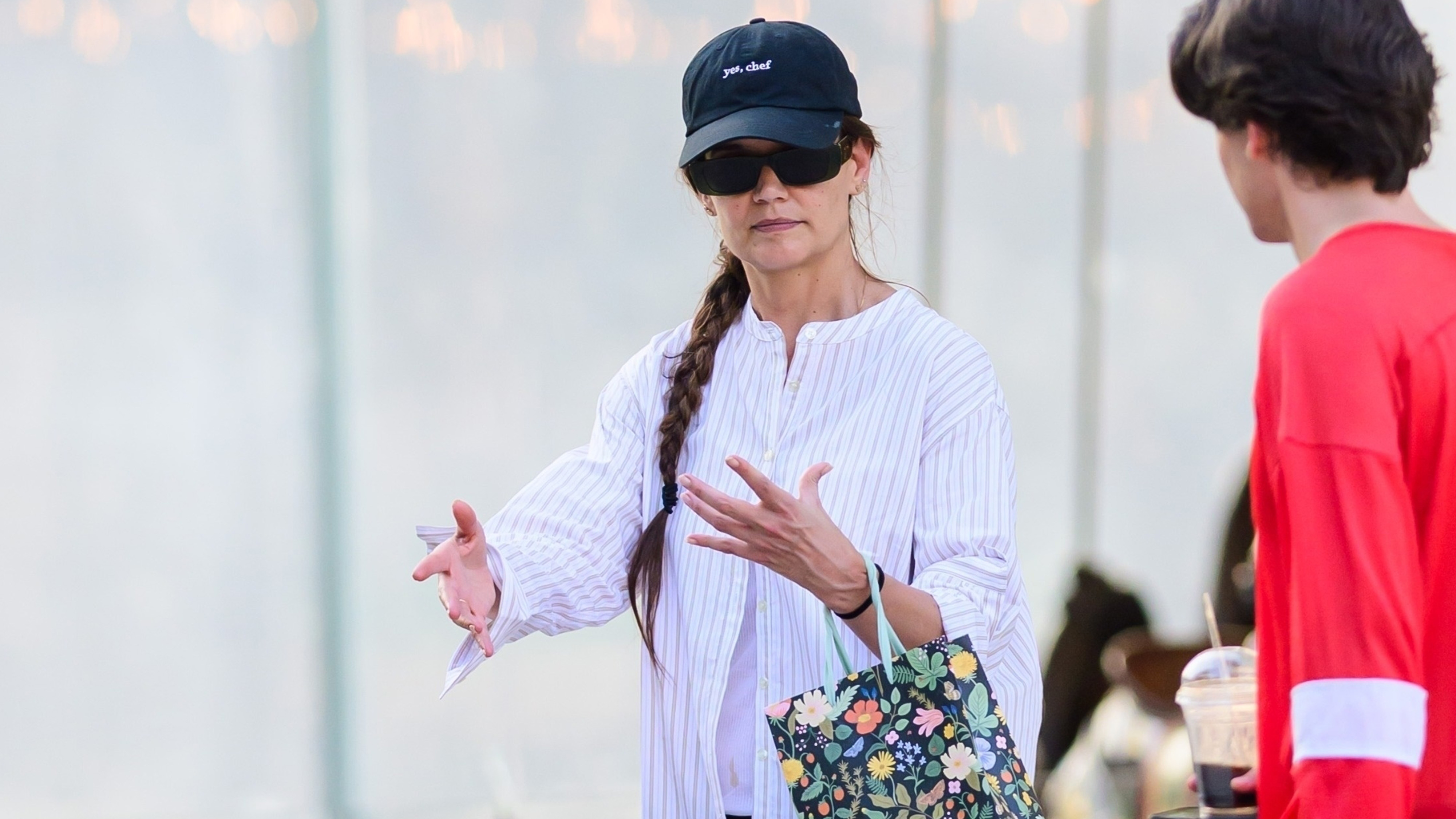Katie Holmes wears a baseball cap, button-down shirt, black shorts, a floral bag, and brown leather loafers.