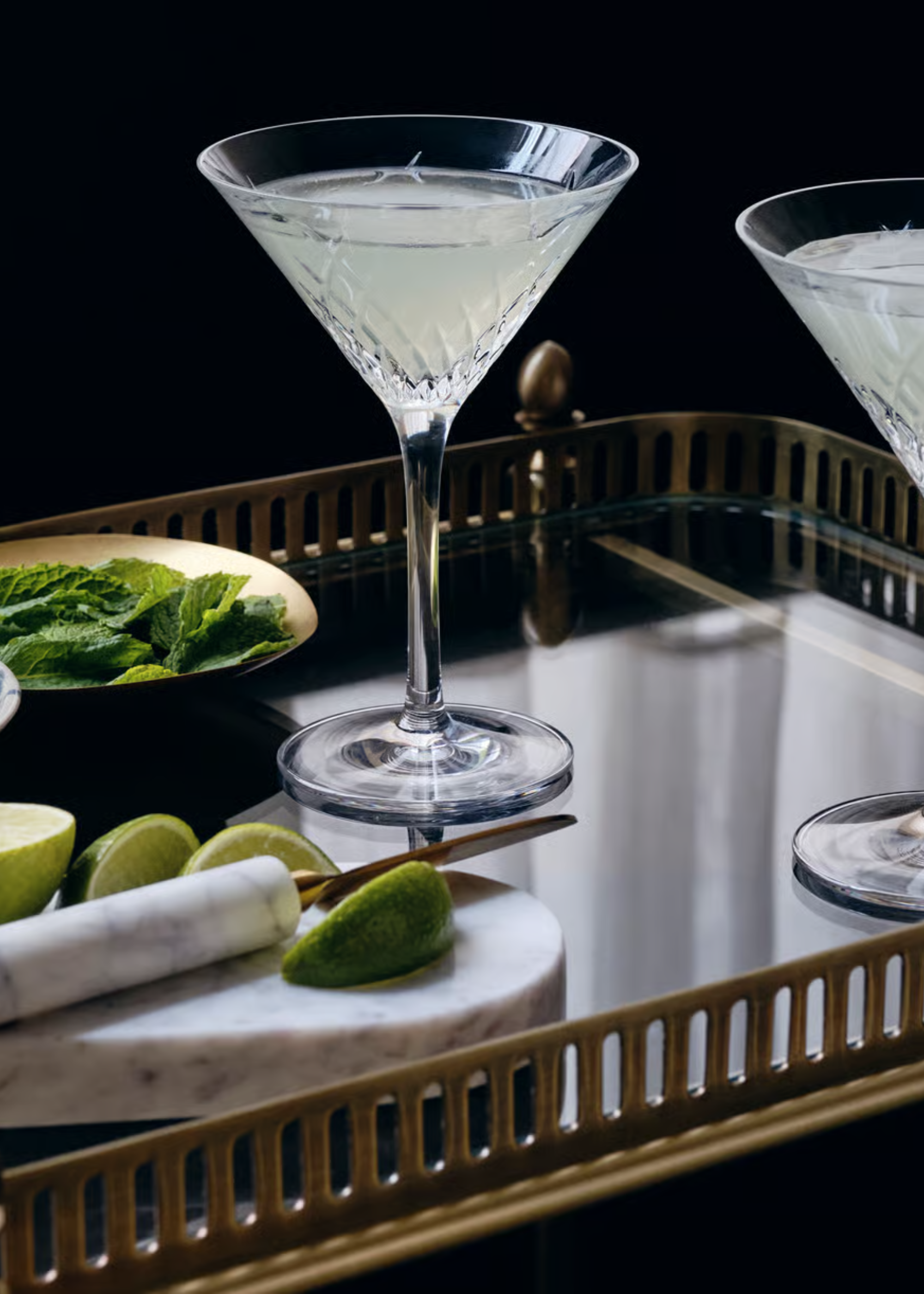 A crystal martini glass on a mirrored tray with a marble knife and chopping board, halved limes, and a bowl of mint