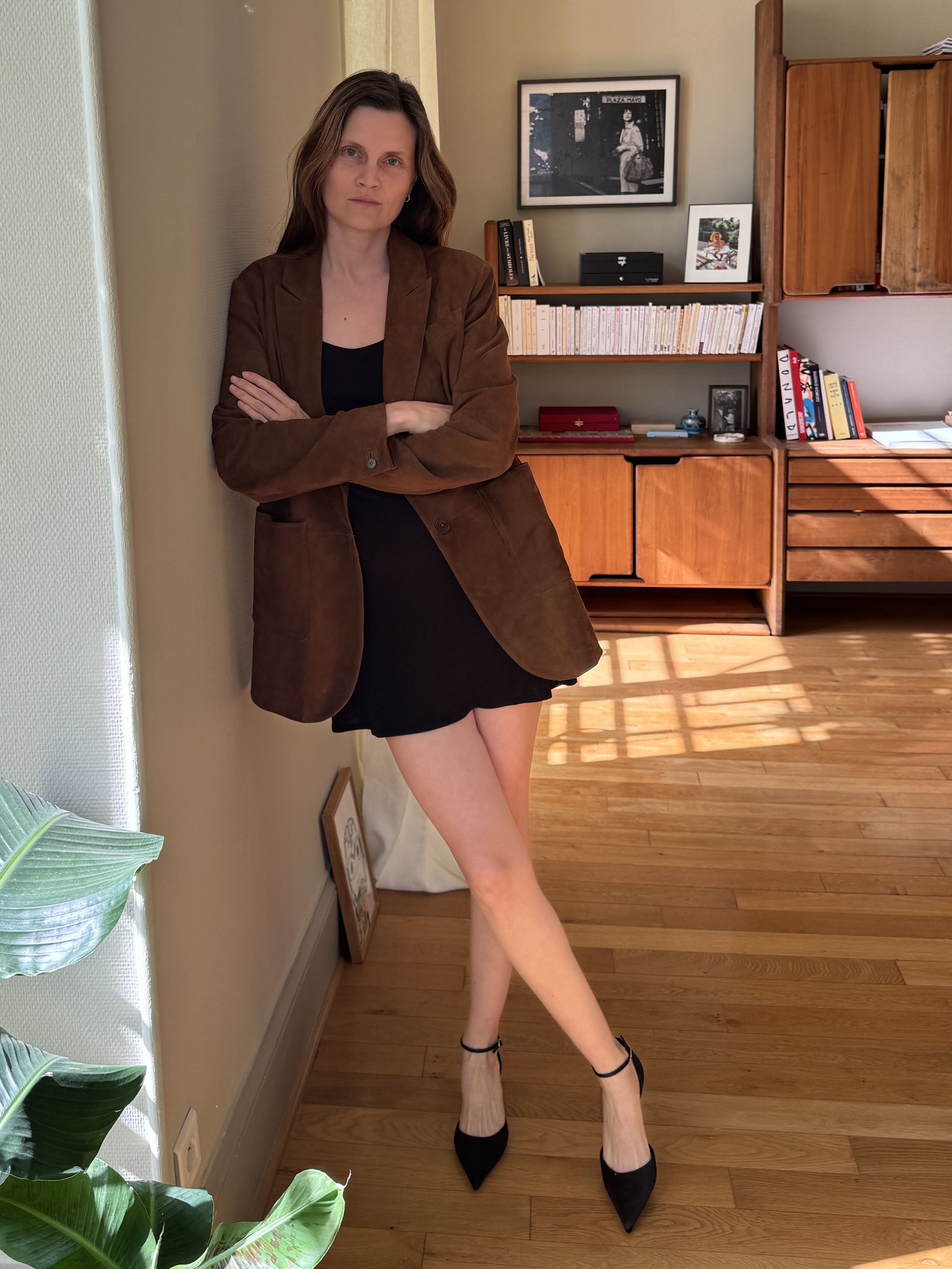 French writer and editor Eugenie Trochu poses in her Paris apartment wearing a brown suede leather blazer, black mini dress, and black ankle-strap pumps
