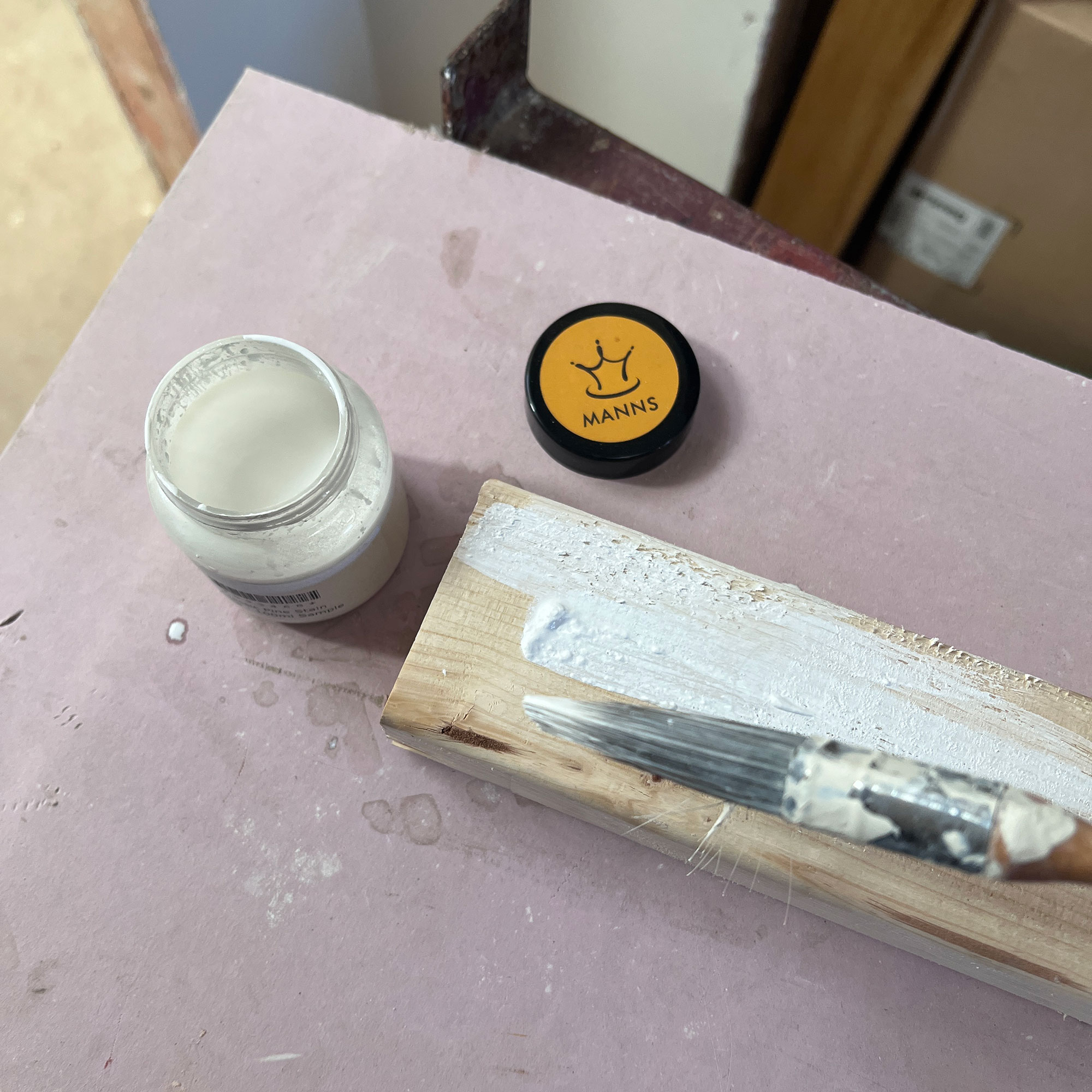 Bottle of white wood stain open on surface beside plank of wood with some painted on