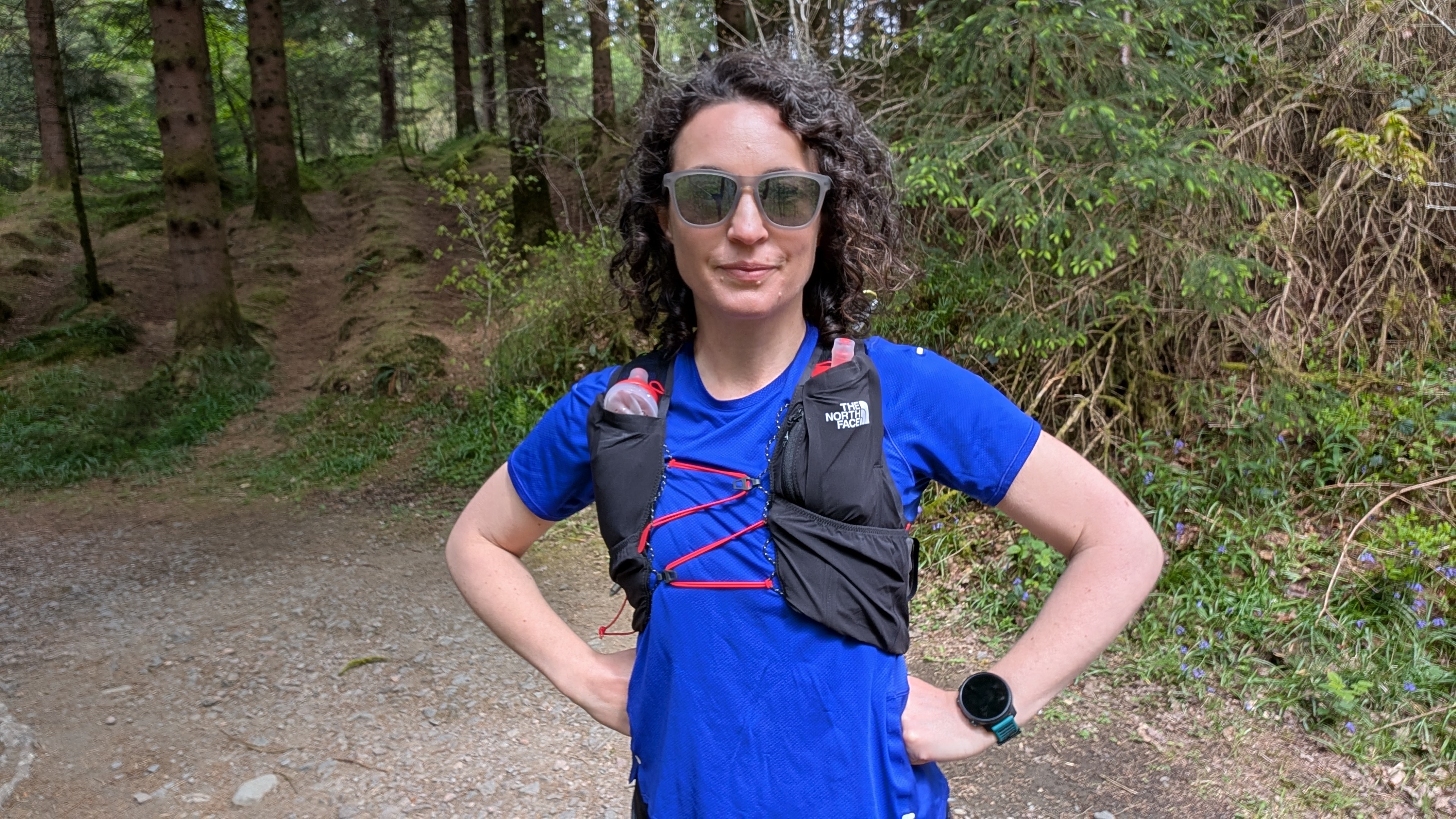 A woman trail runner in the woods wearing a blue t-shirt and a black hydration vest from The North Face