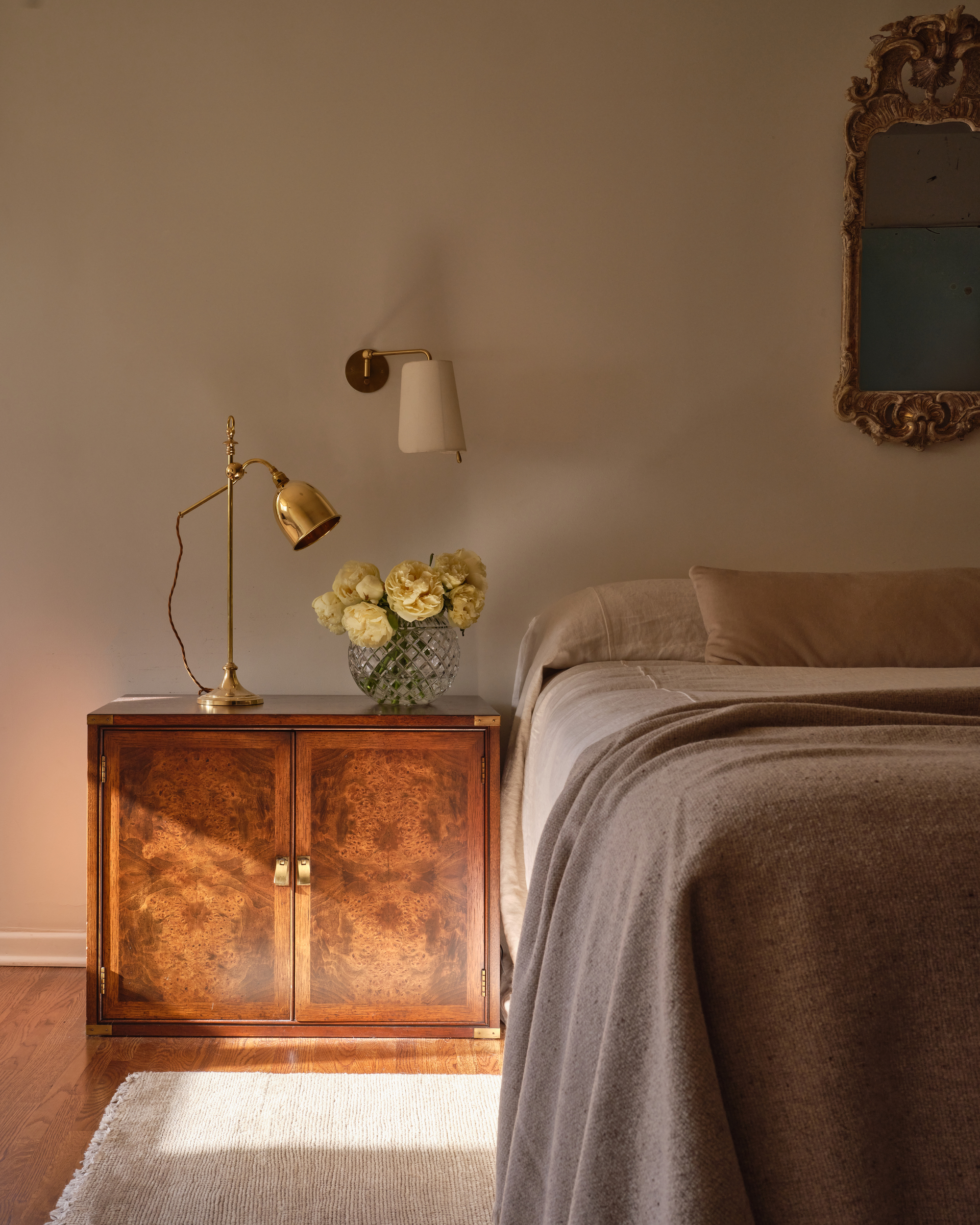 Image of a neutral bedroom with taupe walls and minimalist taupe bedding. There is a vintage-looking wooden bedside table with a gold lamp and a vase of yellow roses.