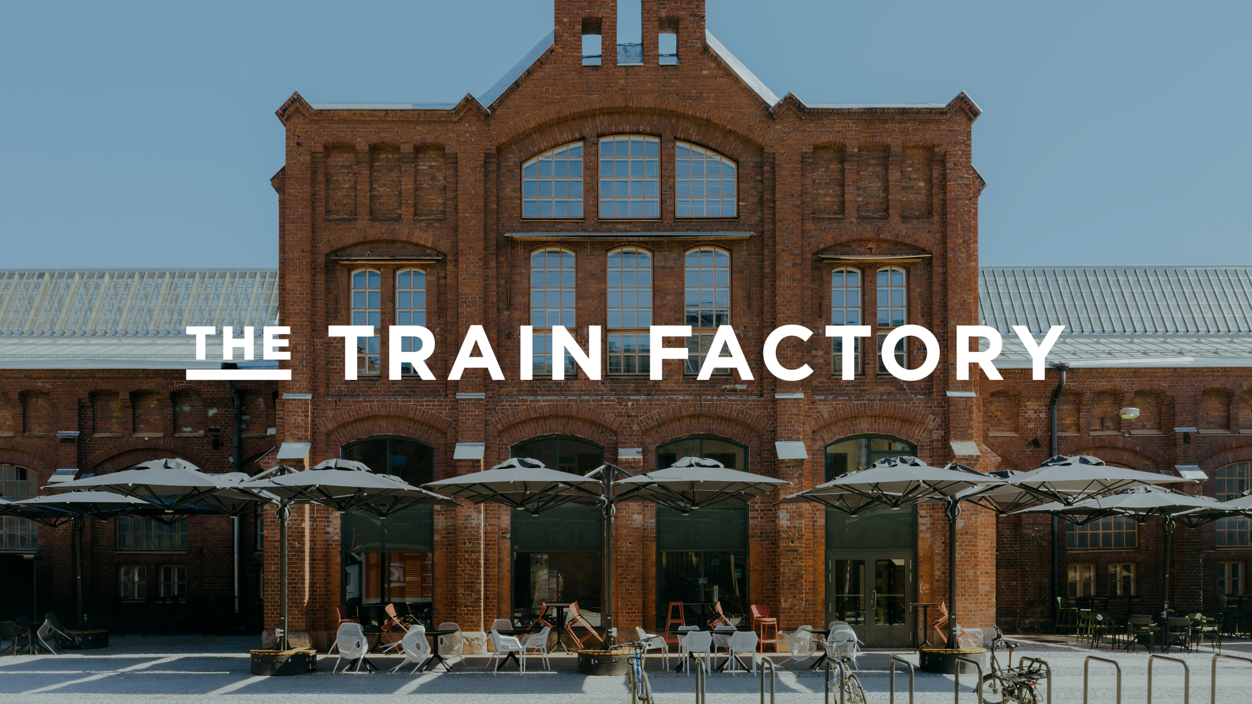 A red brick former train factory building with tall arched windows and rows of grey umbrellas shading outdoor seating, with "The Train Factory" logo overlaid in white.