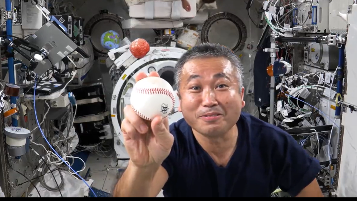 astronaut holding a ball in front of a camera with a grey space station module in behind, crowded with experiments