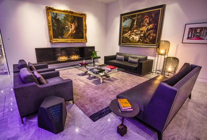 A lounge area with a purple leather sofa, purple fabric chairs, a black leather sofa, a fireplace, floor lamps, coffee tables, a glass table and large wall paintings.