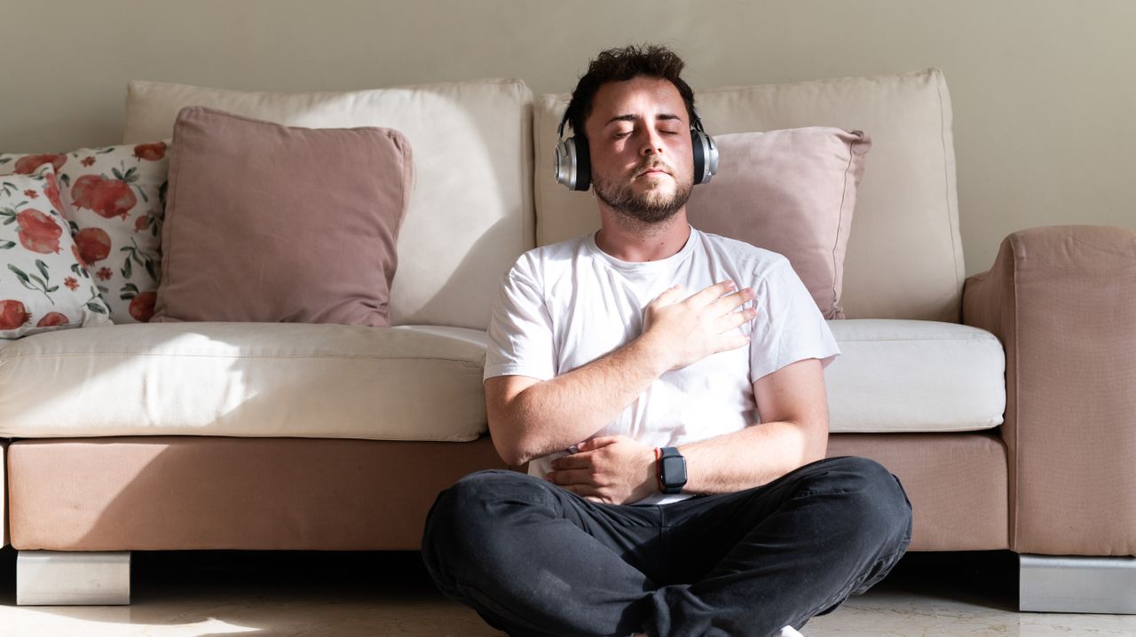 A man sits cross legged on the floor in front of a couch with his eyes closed. One hand rests on his stomach, while the other rests on his chest. He wears a t-shirts and sweatpants and he has on a set of over-ear headphones. 