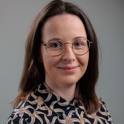 A close up of a woman with long brown hair wearing a patterned shirt and glasses who smiles at the camera