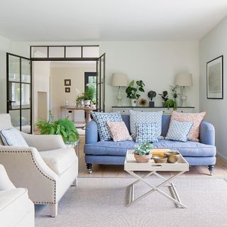 living room off the entrance hall with a blue couch, cream armchairs and a grey rug, with a foldable wooden tray coffee table in the middle, with pot plants dotted around the room