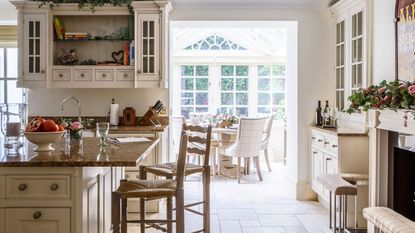 A white festive kitchen with white cabinets, a marble kitchen island, tiled floor, two wooden bar stools, a large fireplace to the right with a festive garland, and a bright, airy and spacious dining room seen behind with large windows out onto a leafy garden.