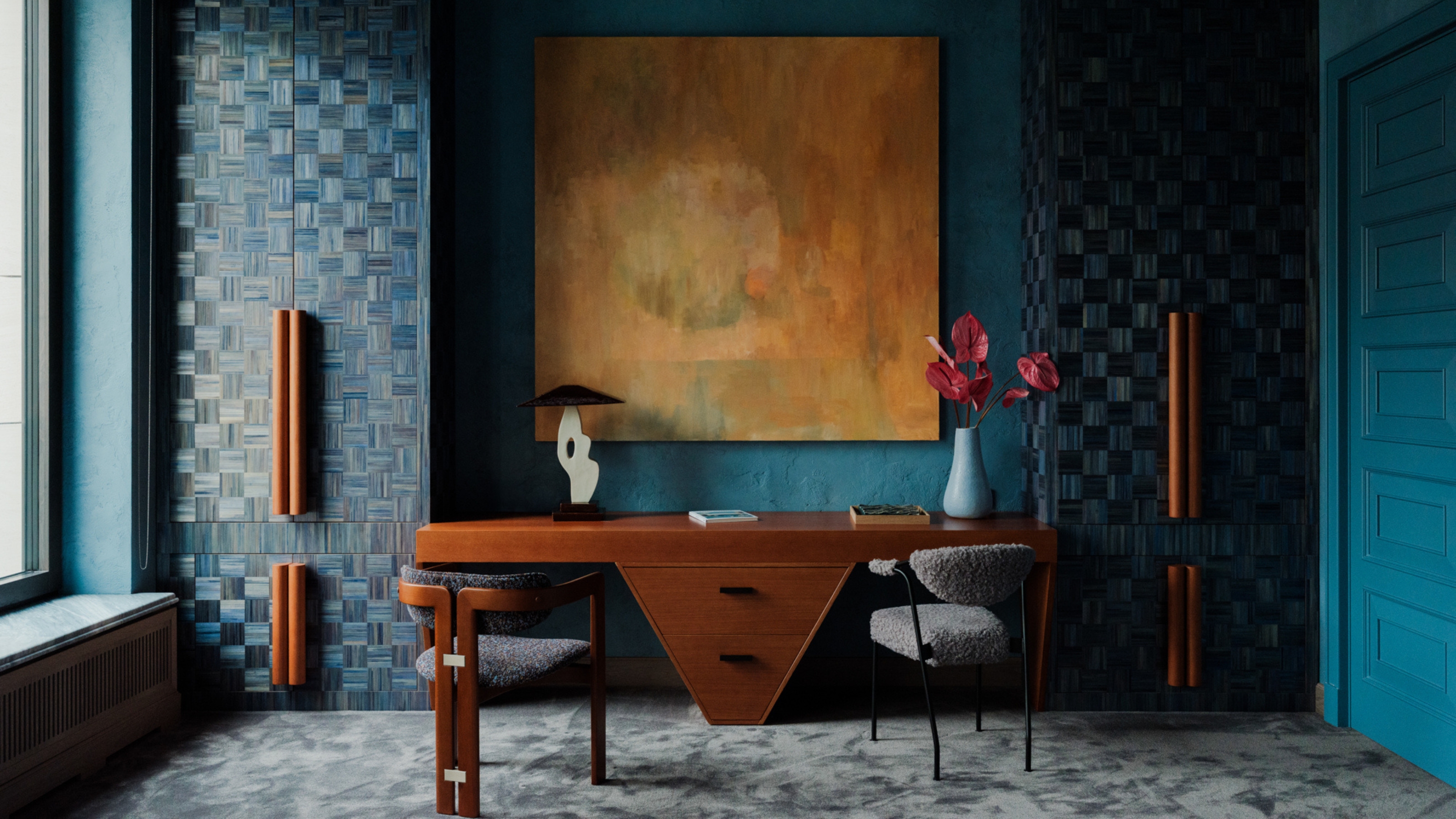 Image of a bright, teal home office with a luxurious aesthetic. There are floor-to-ceiling cabinets in a woven, monochrome teal design. With large orange handles. There is a large orange painting over the wooden desk with a triangular bottom. 