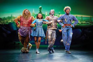 four people (kyle ramar freeman as lion, nichelle lewis as dorothy, phillip johnson richardson as tin man, avery wilson as scarecrow) stand with their arms linked in front of an emerald city background