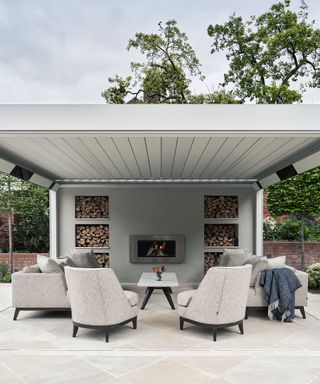 A stylish outdoor living area with upholstered chairs and sofas and an outdoor fireplace built into a wall with log stores