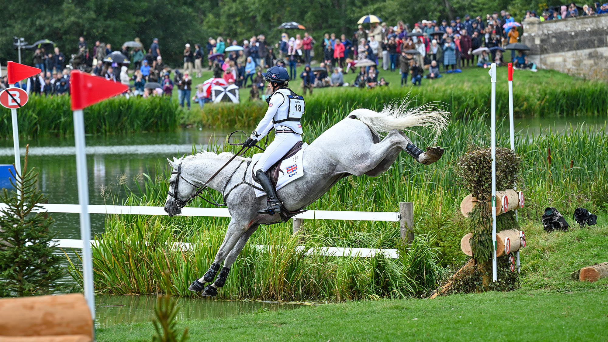 Piggy March, a British rider in a white top and blue hat silk, rides grey horse Halo in a flying leap over a narrow brush fence into water on the Blenheim cross-country course