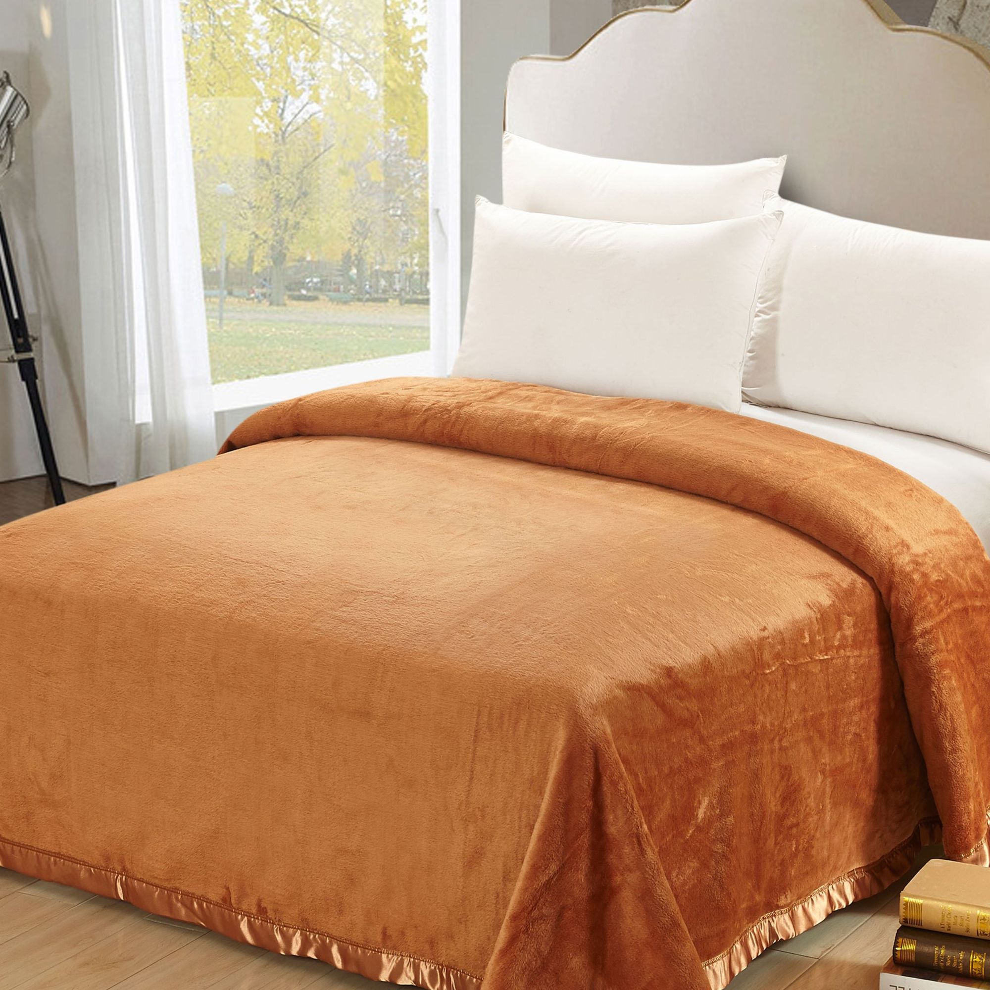  Orange silk blanket on made bed with white pillows and white headboard, in front of window revealing green outdoor space.