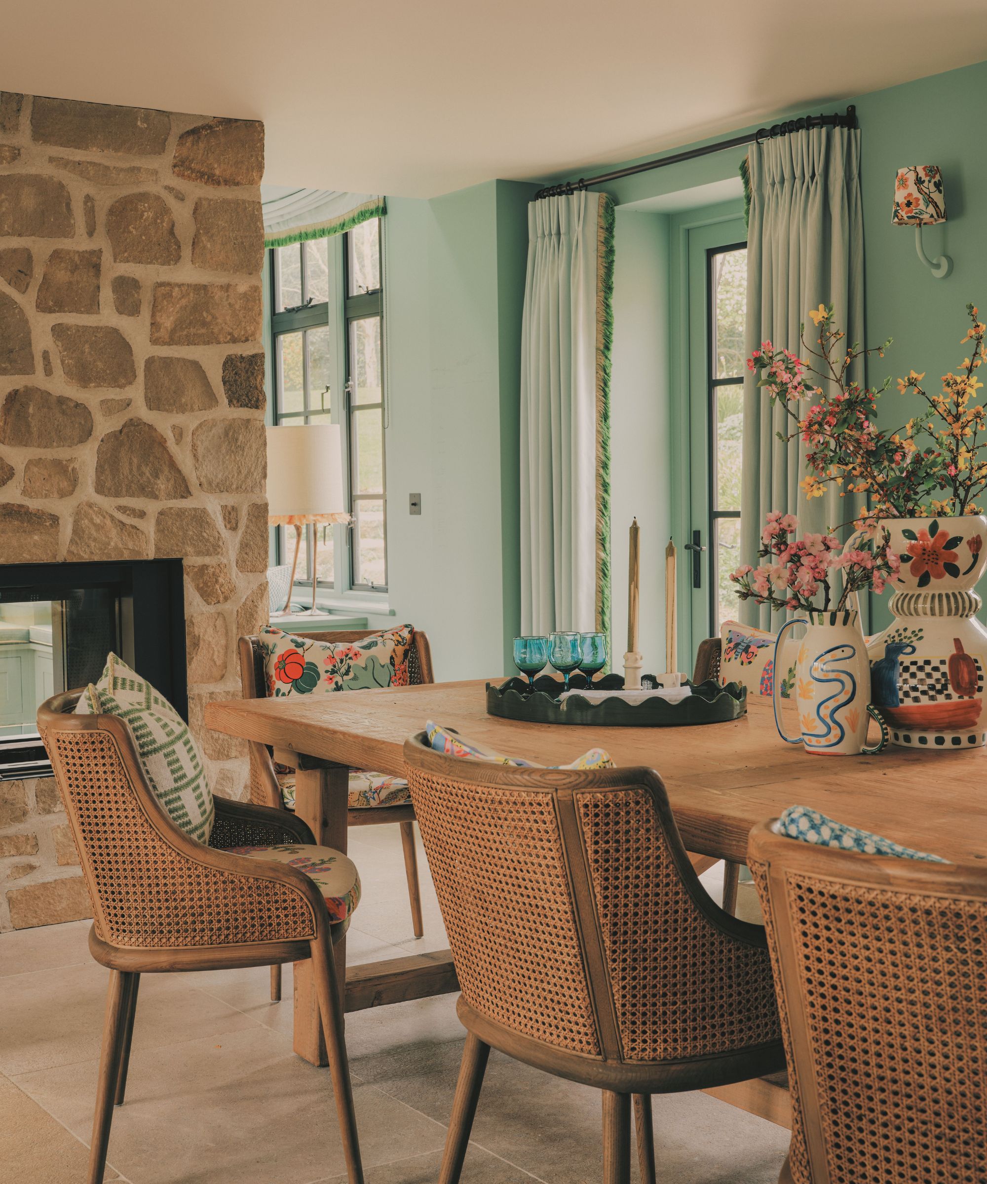 Rustic dining room with woven rattan chairs around a wooden table, mint green walls, a stone fireplace, and decorative floral ceramics.