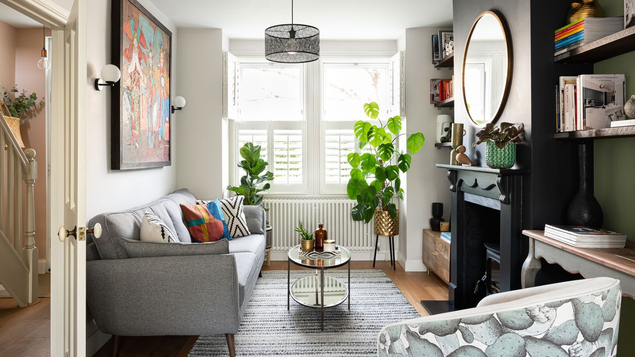 Living room with wooden floors, white left-hand wall and green right-hand wall, black painted fireplace, round mirror, glass coffee table, grey sofa, pattern armchair and blinds in window