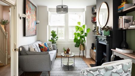 Living room with wooden floors, white left-hand wall and green right-hand wall, black painted fireplace, round mirror, glass coffee table, grey sofa, pattern armchair and blinds in window