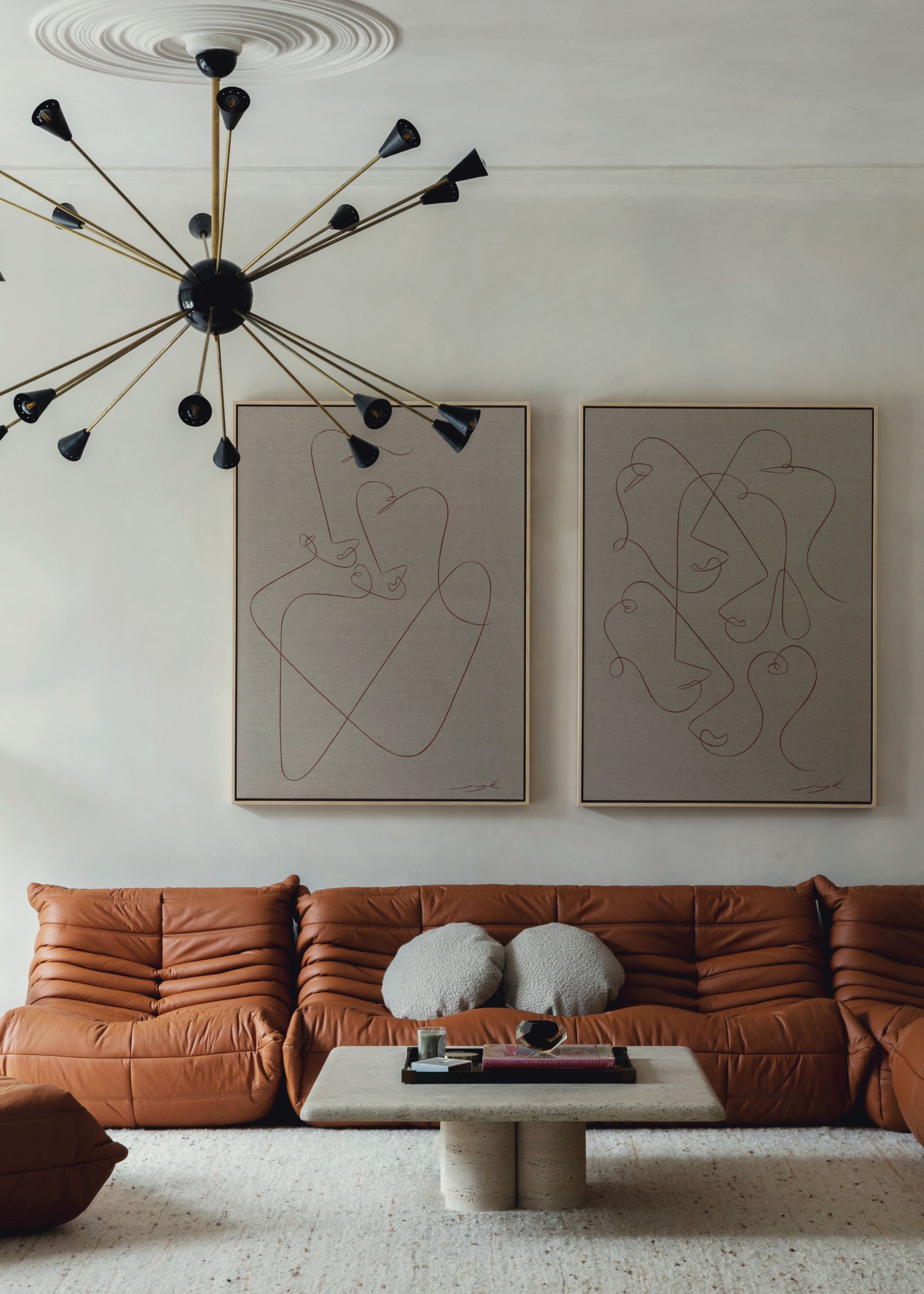 A modern living room with pale gray walls, a carpeted floor, and a large tan leather slouchy sofa against the wall with two minimalist artwork pieces on the wall.