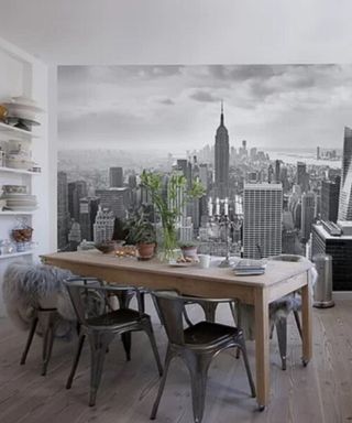 A gray dining room with a black and white mural of a city, a dark brown wooden table with plants and tableware on it, black metal chairs underneath it, and wall shelves to the left