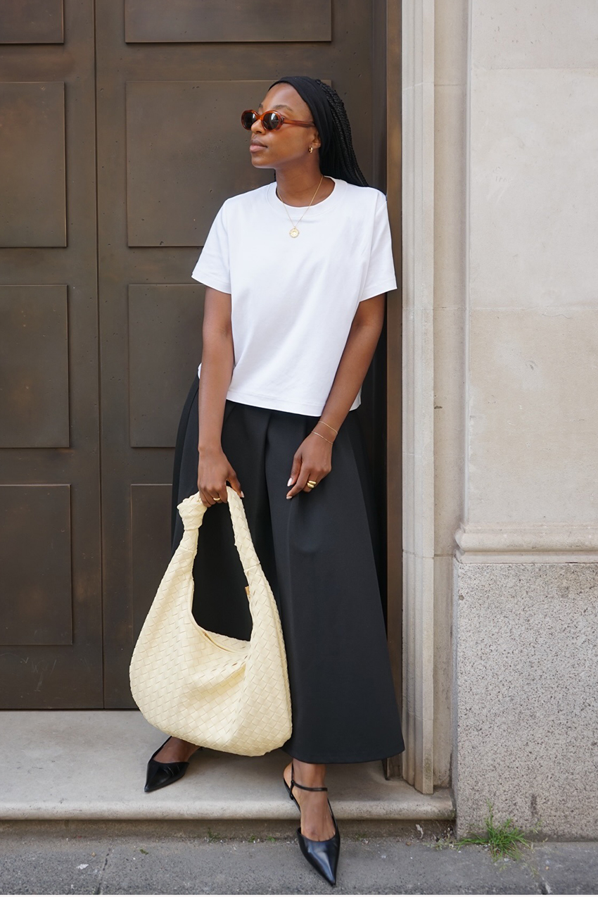 a woman wearing a white cropped tee, black culottes, black pointed-toe shoes, and a white woven bag