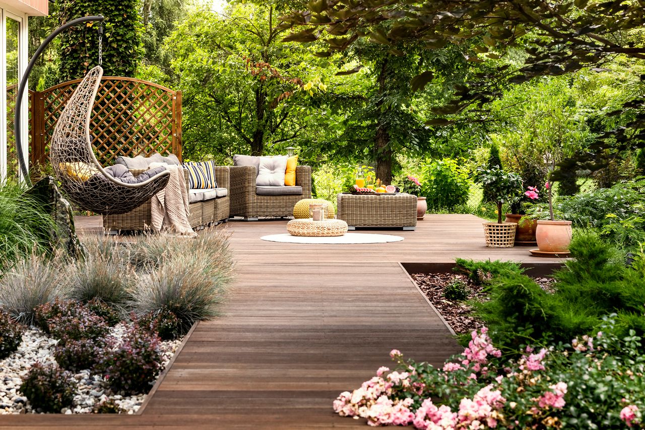 a stunning deck in a green garden with lots of huge trees, full of wooden garden furniture including a hanging chair and arm chair