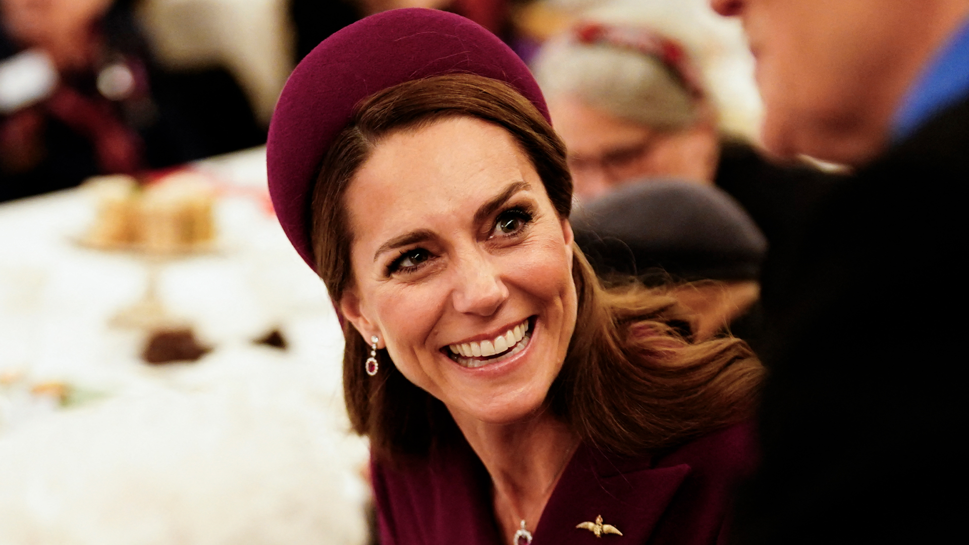 Kate Middleton wears a wine red coat dress and matching hat and laughs
