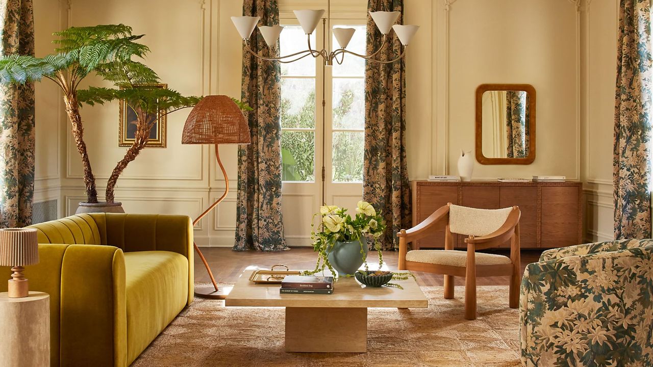 a living room decorated with lulu and georgia&#039;s spring collection - a jute rug, travertine coffee table, yellow velvet sofa, verdure printed armchair and curtains, a pendant light, and plant in the corner