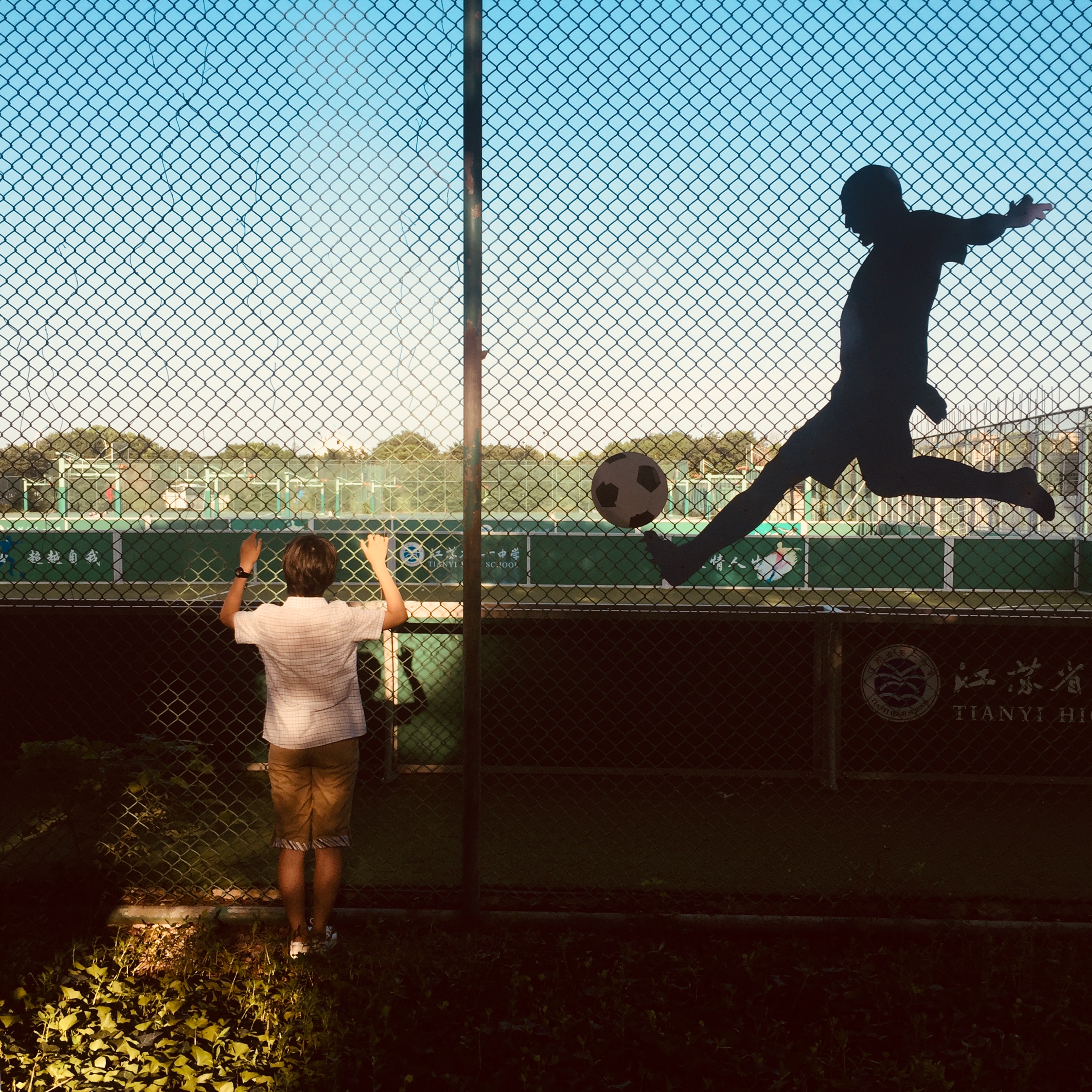 A silhouette of a man kicking a ball while a child watches from behind a fence