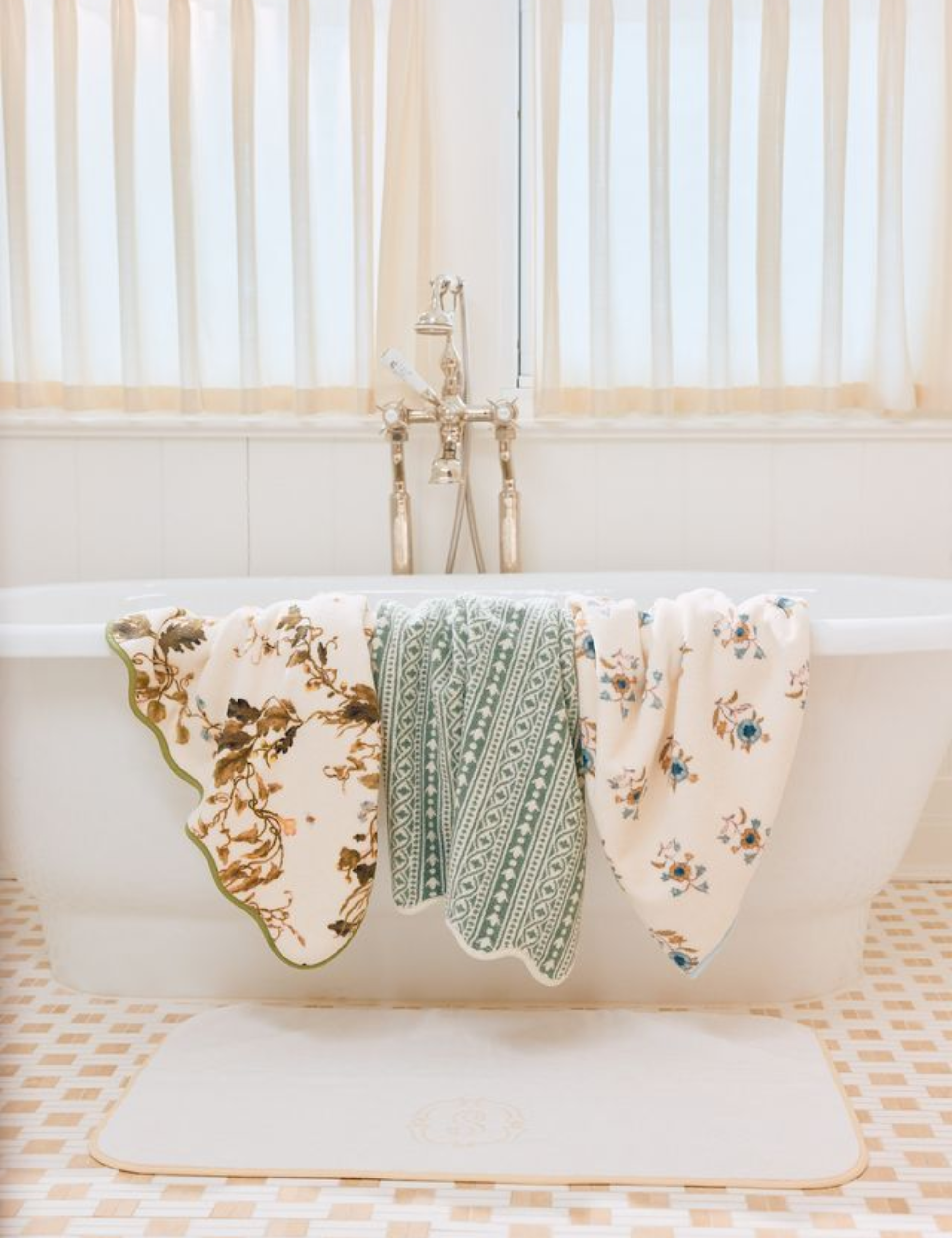 Airy tonal cream bathtub vignette styled with sheer curtains and three floral, scalloped-edged bath towels in various prints.